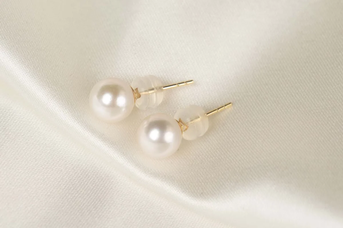 Two white pearl stud earrings with gold-colored posts on ivory satin.
