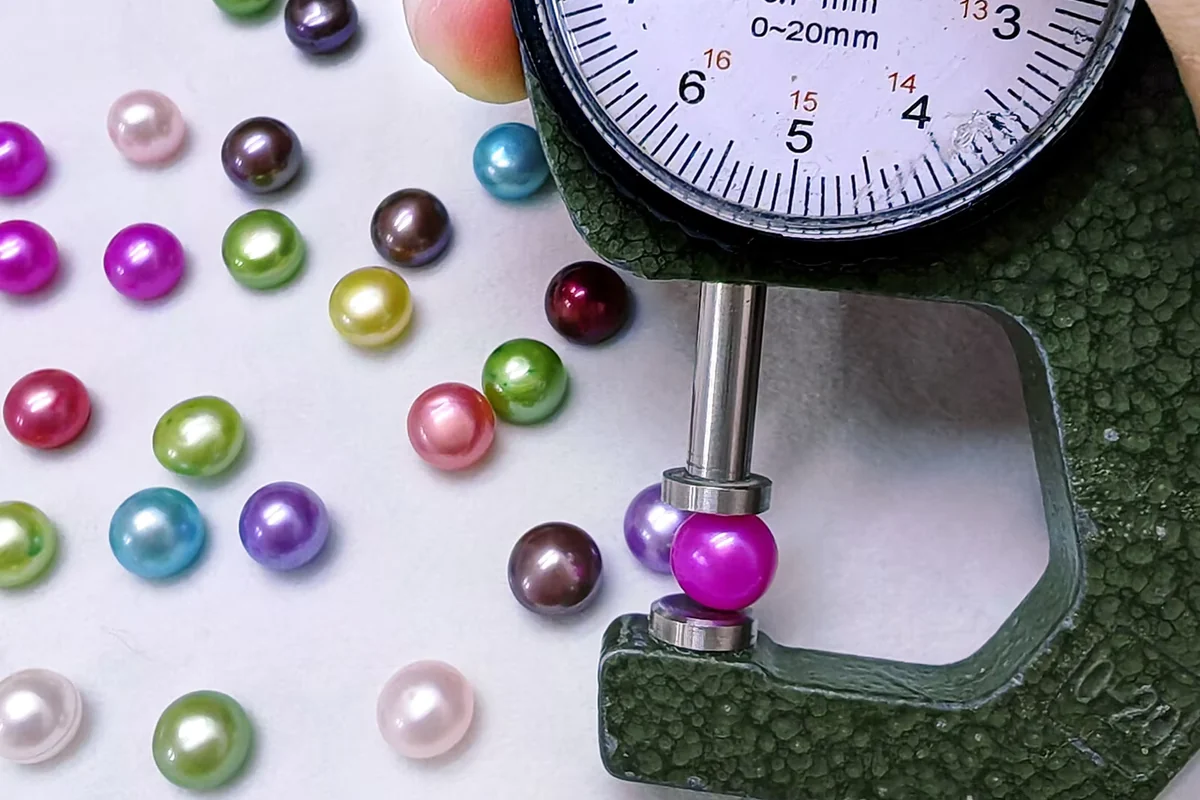 A dial gauge measuring a colored pearl beside loose pearls.